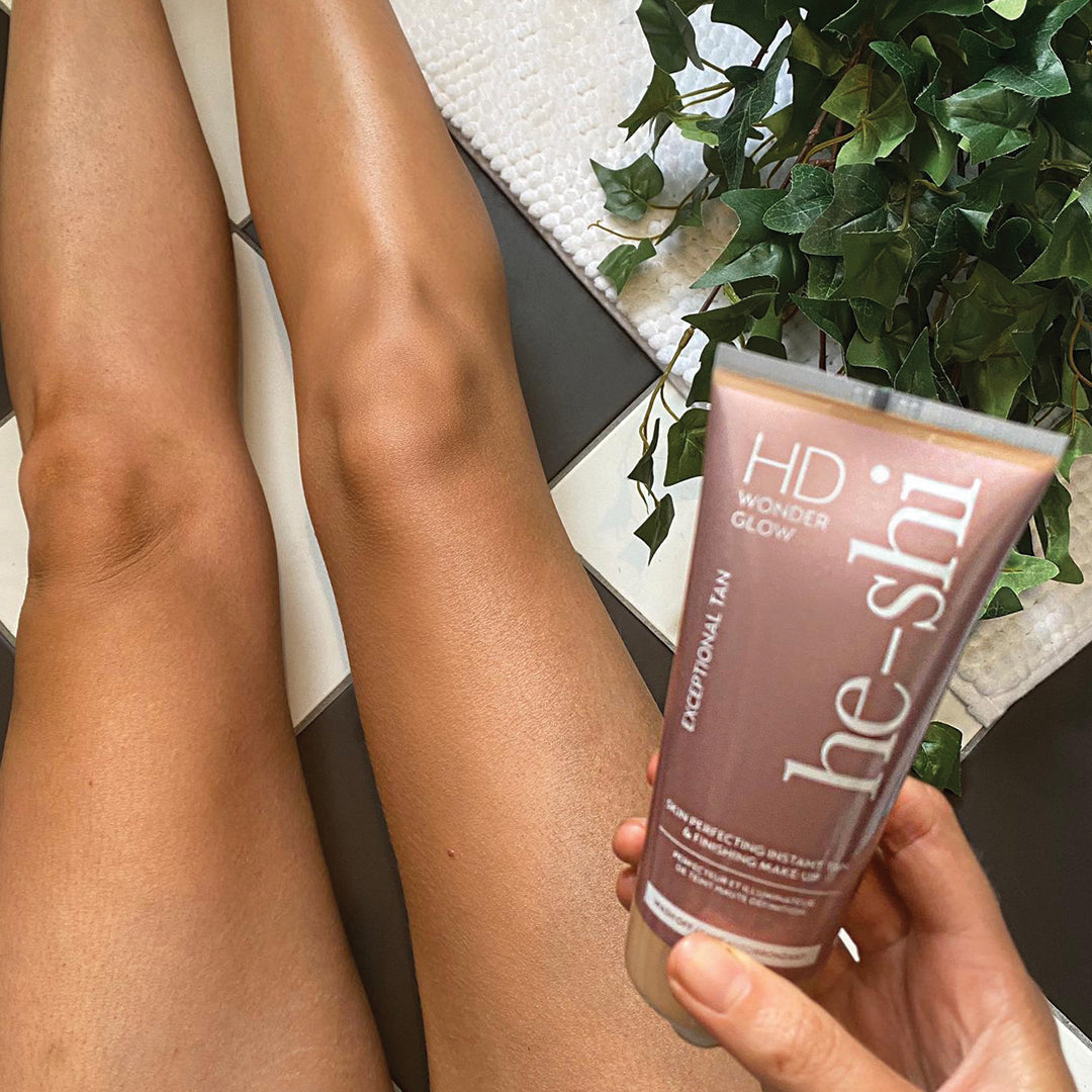 Image of woman sitting on bathroom floor holding a tube of He-Shi HD Wonder Glow. The image shows the woman's legs only to demonstrate the natural looking tan with an irredesant finish.